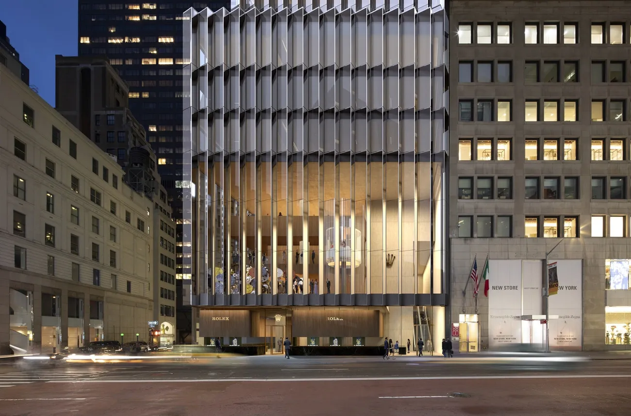 Night view of the illuminated Rolex Building by David Chipperfield Architects with vertical glass fins and street traffic.